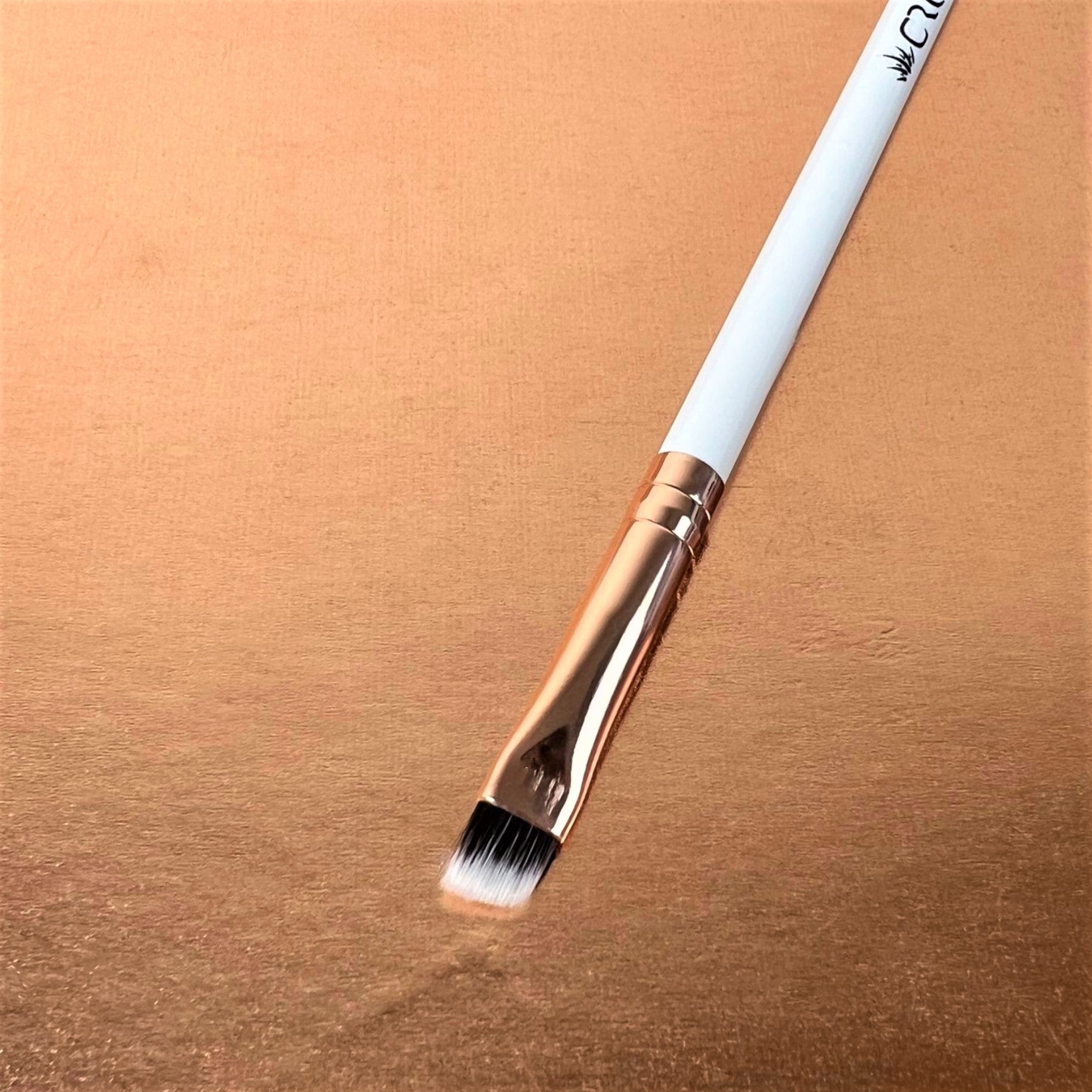 A single angle liner makeup brush with a white handle and gold ferrule, showcasing the brush head resting on a rose gold-colored surface.